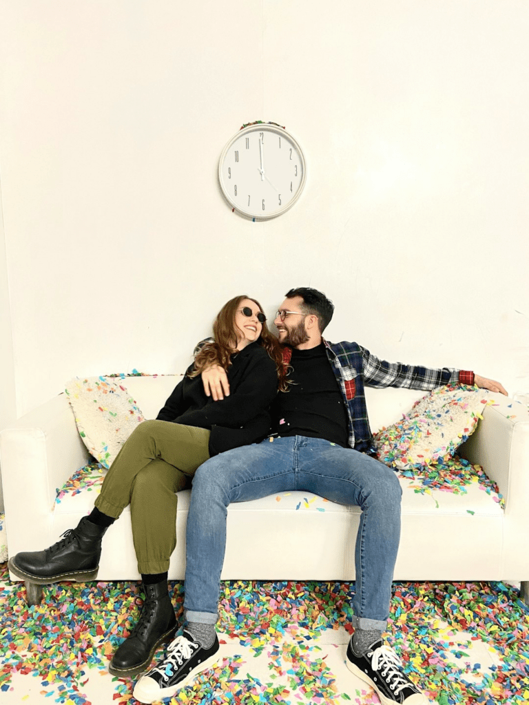 clyjch5i Junges Paar sitzt auf einer Couch, die mit Konfetti bedeckt ist. Das Paar sieht sich and und lächelt dabei. Der Mann hat seine rechts Hand auf der Schulter der Frau.